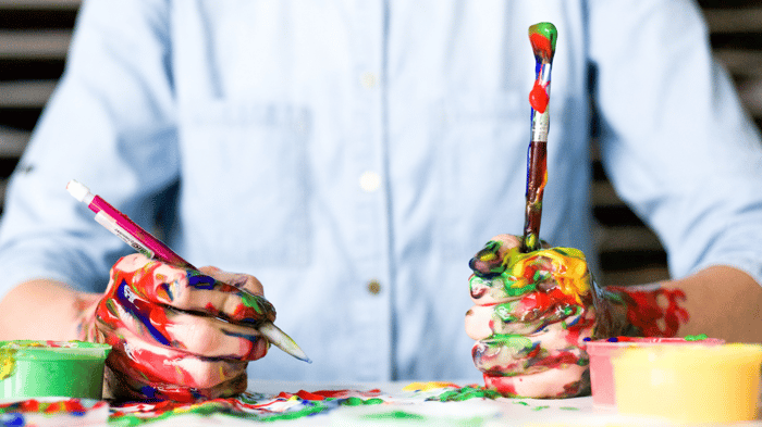 A close shot of a man in a buttoned up office shirt holding a pen in one hand and a paint brush in the other and both, including his hands, are covered in messy multi-colored paint