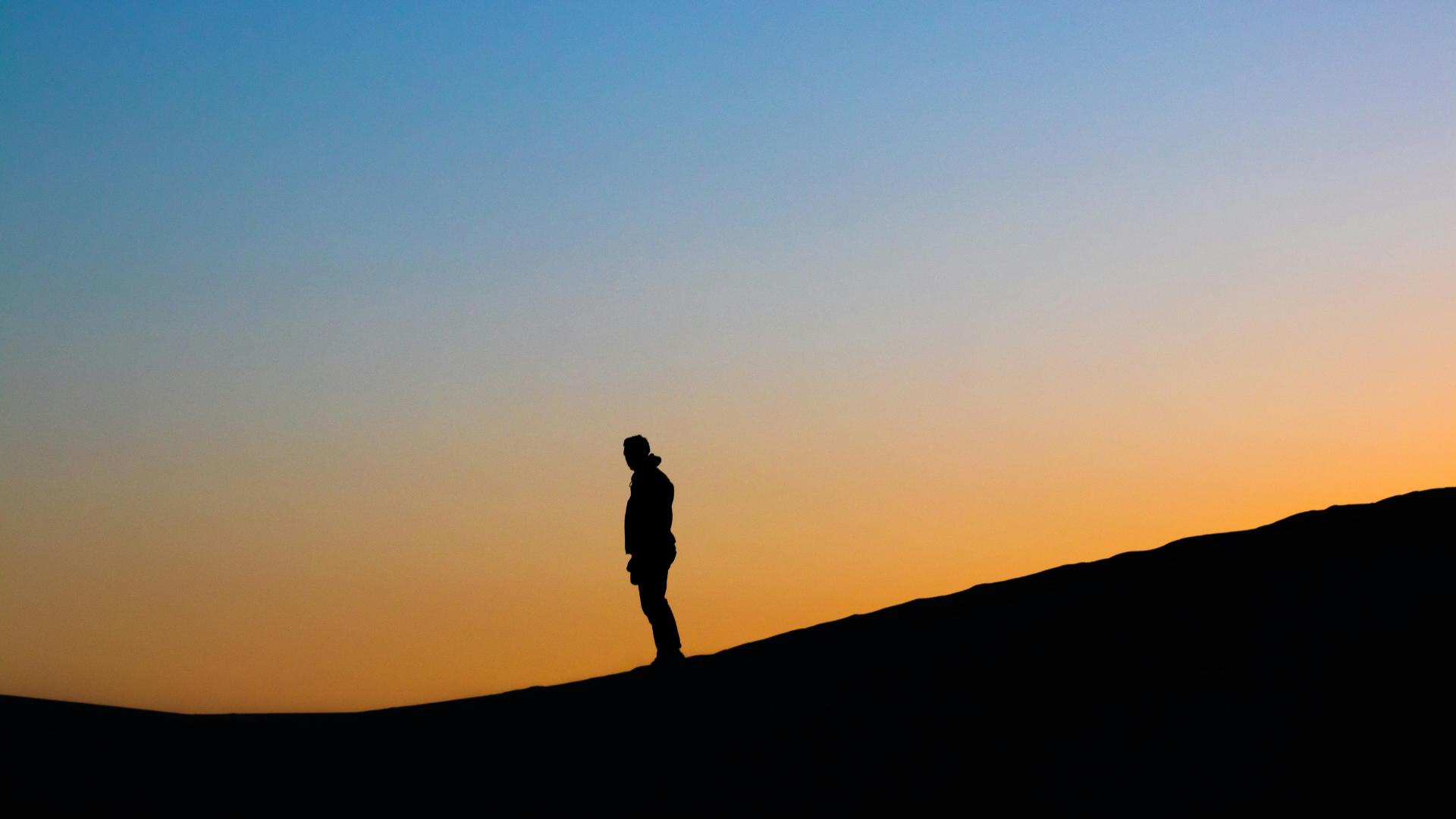 The dark silhouette of man's figure standing a top a sloping during golden hour, a warm rusty orange sunset in the background
