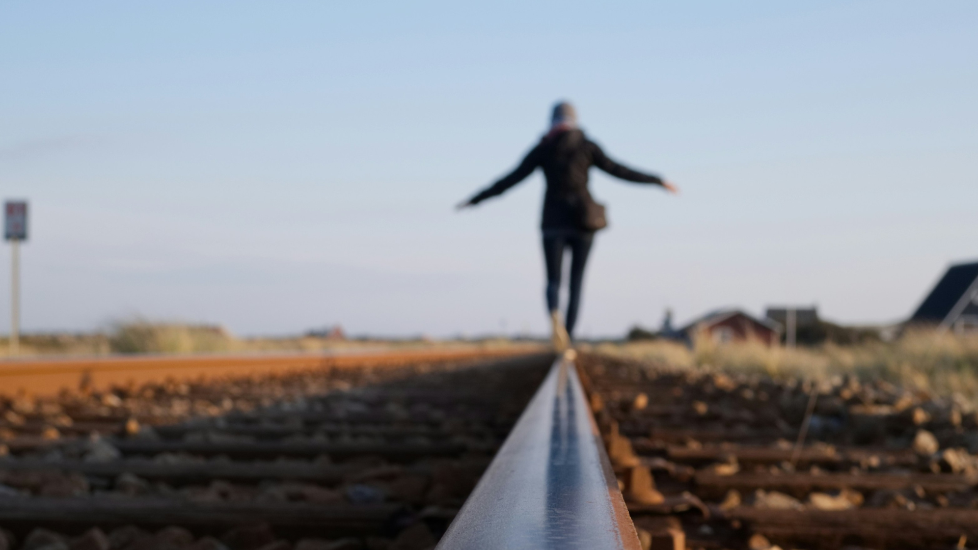 A slightly out of focus figure of a person in the distance with arms stretched out left and right to balance while walking along a single steel rail of a railroad track