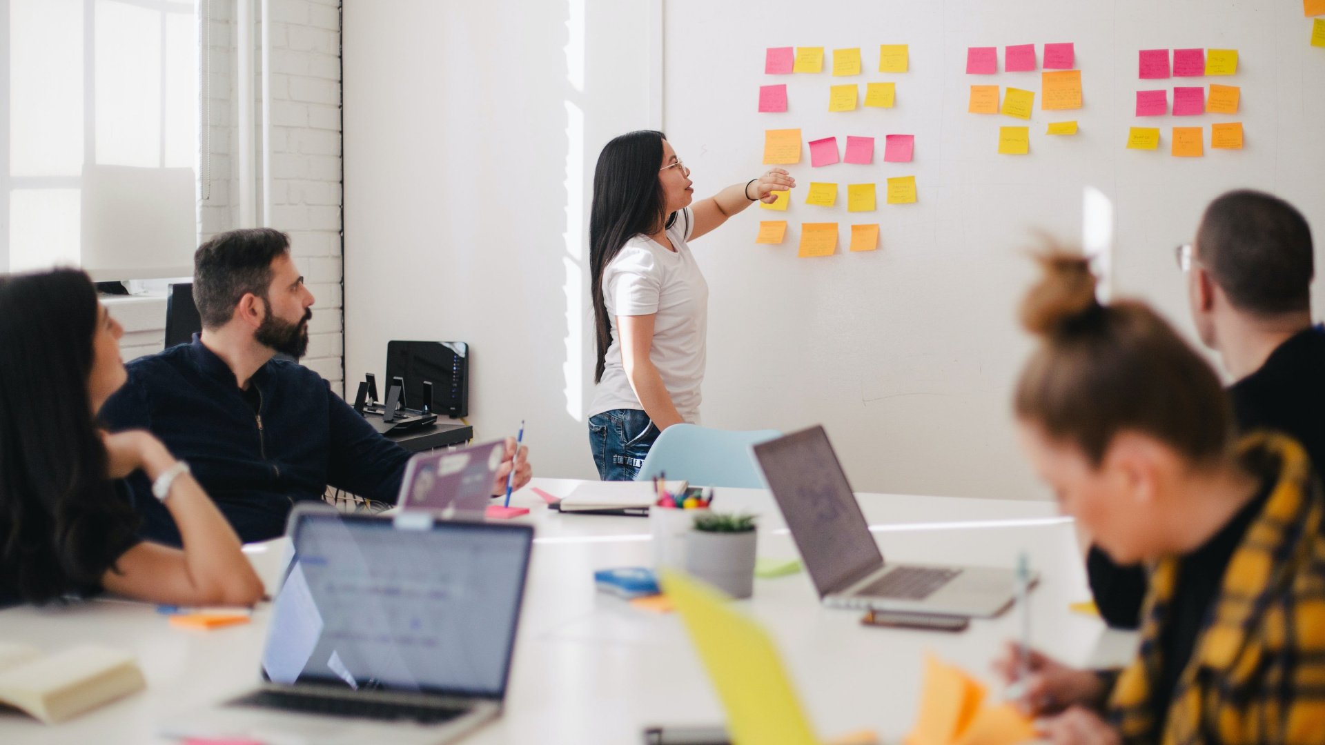 A group of people in a meeting, sitting at a table and looking at a white board with sticky notes on it