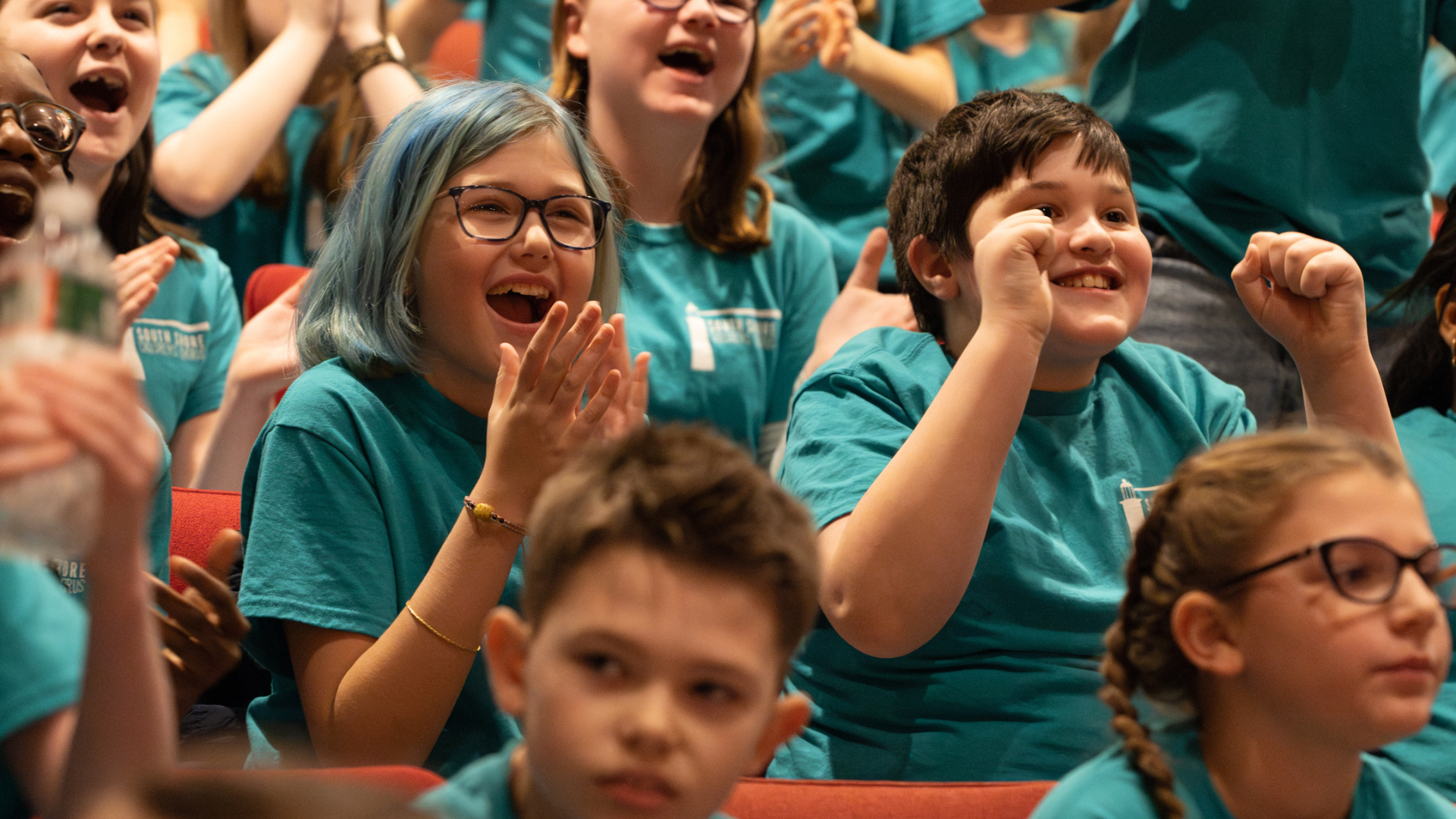 A group of youth wearing matching green T-shirts laugh, smile and cheer as they sit in an audience