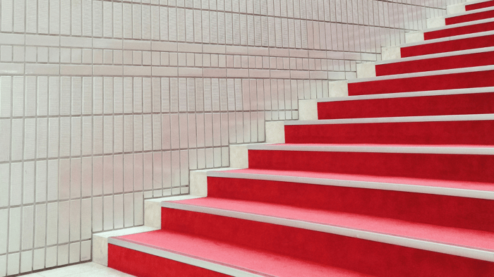 Vibrant red stairs stand out against a white tiled wall as they ascend toward the top right corner of the image