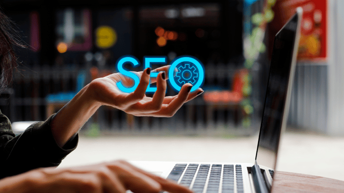A person is working on a laptop in an office, with one hand on the mouse and the other raised, appearing to hold a floating, glowing blue "SEO" text with a gear icon. The background is blurred, featuring colorful lights and a modern setting.
