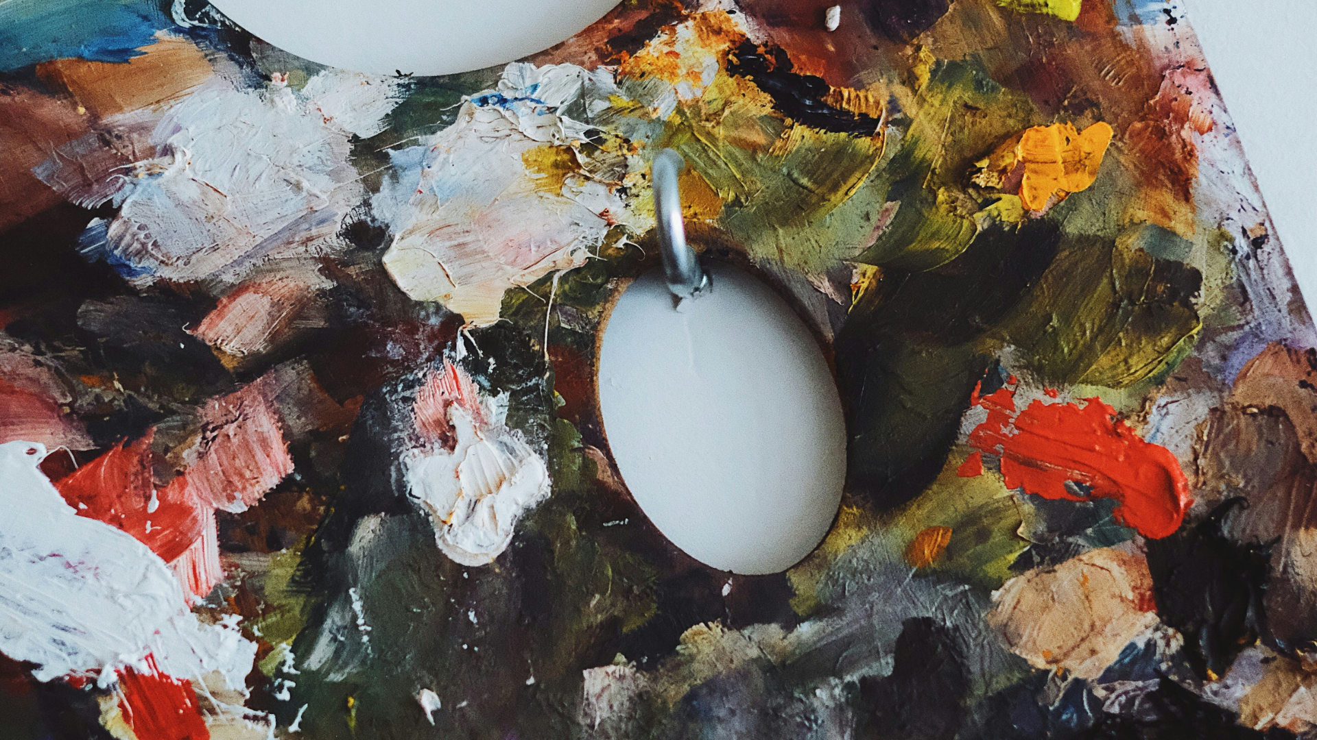 A close photo of an artist's mixing palette covered in painterly and messy strokes of moody dark colors with splashes of red and goldhanging on the wall of an art studio.