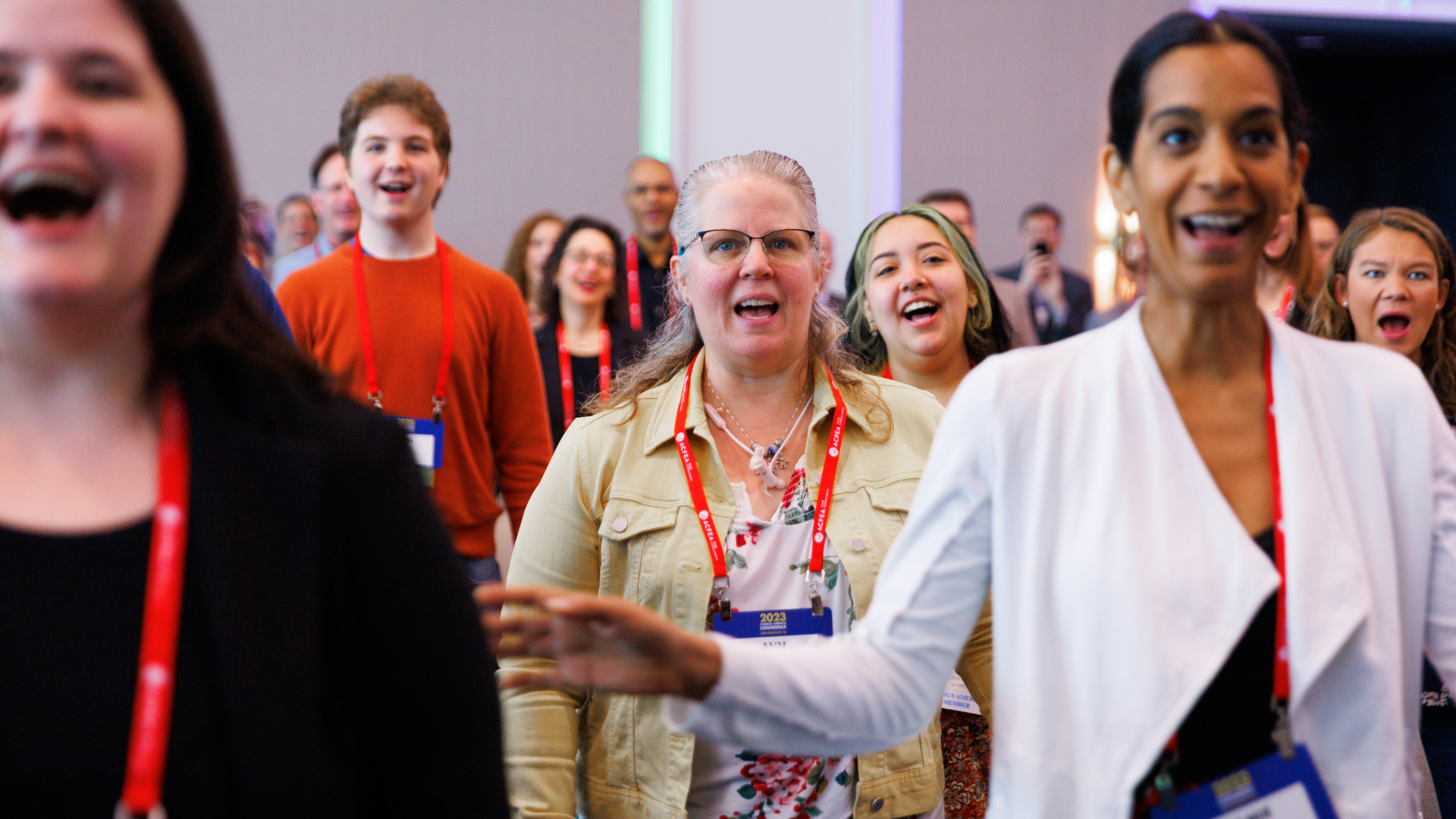 A room full of conference attendees wearing red lanyards and name tags with their mouths open as they face the camera and sing