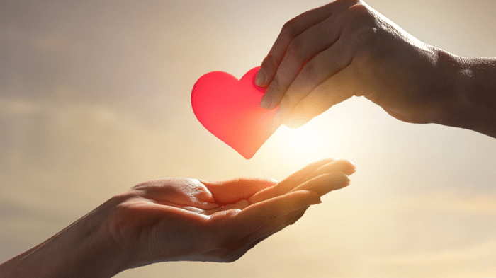 A photo of two hands, one holds a red paper heart above the other, which is cupped below, as if reaching to hold the heart