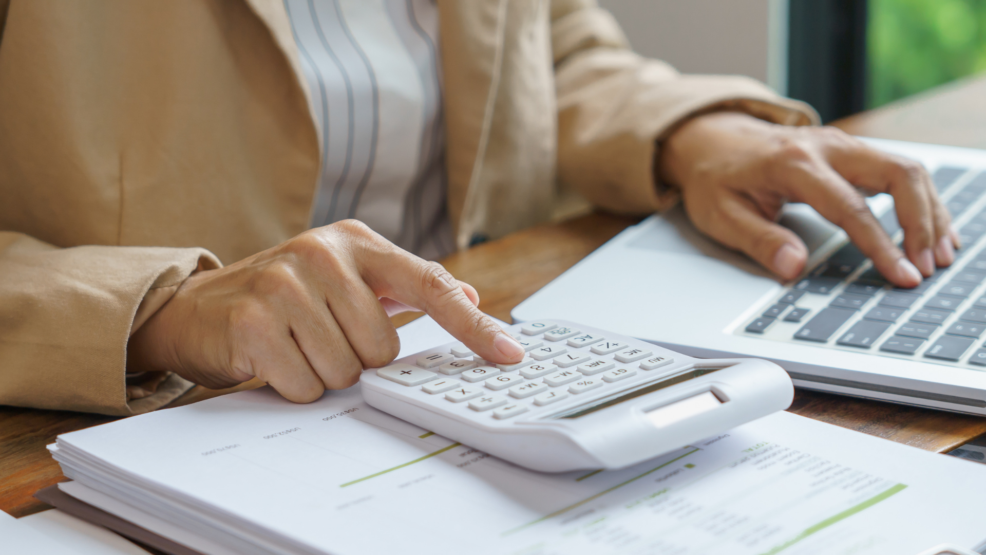 Tightly cropped photo of a person at a desk with one hand on a laptop keyboard and the other hand typing on a calculator