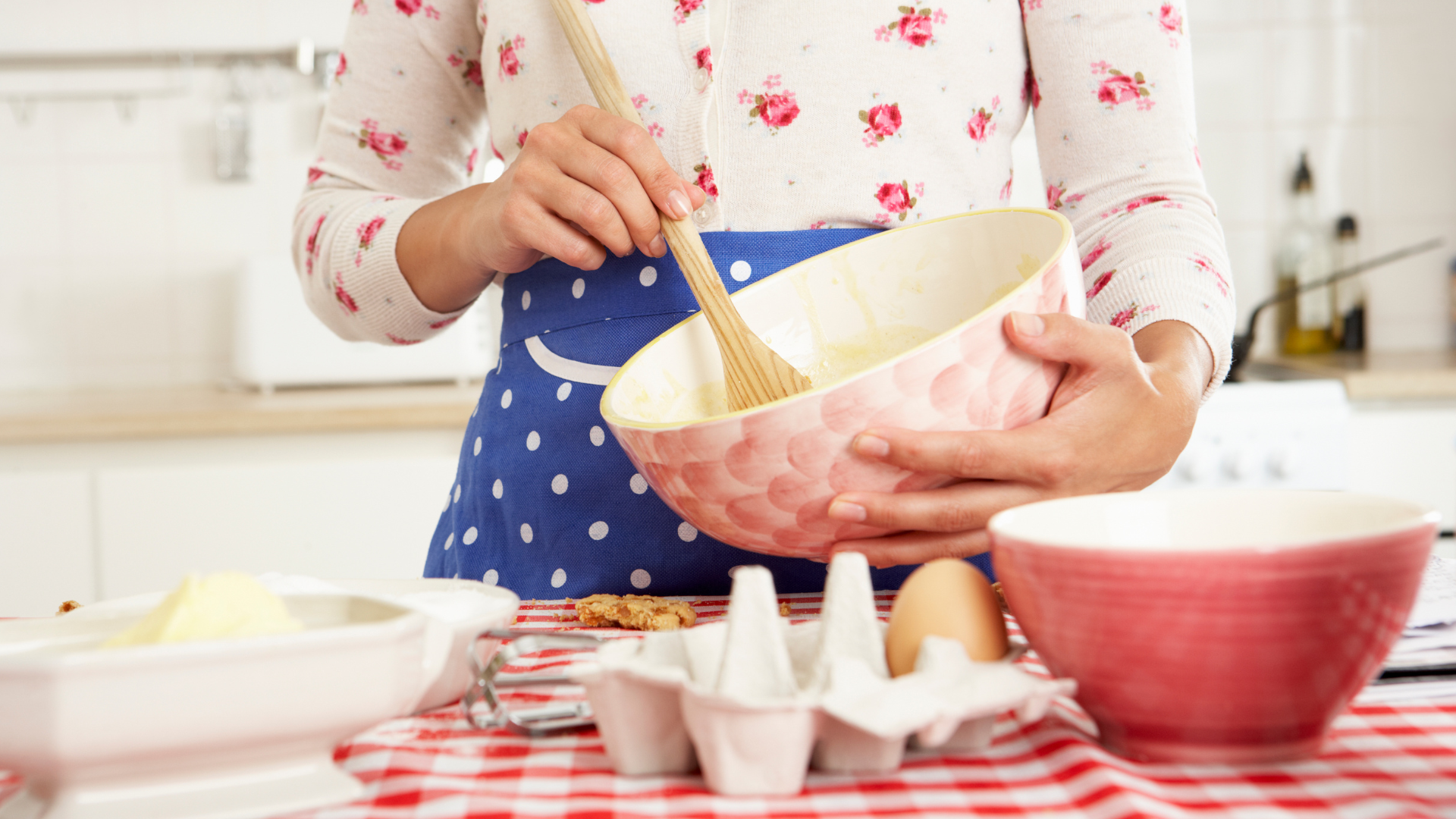 A person stands behind a kitchen counter, fresh eggs and a dirty mixing bowl in view, holding a red mixing bowl while they stir the bowl's contents with a wooden spoon the person is in a kitchen baking.