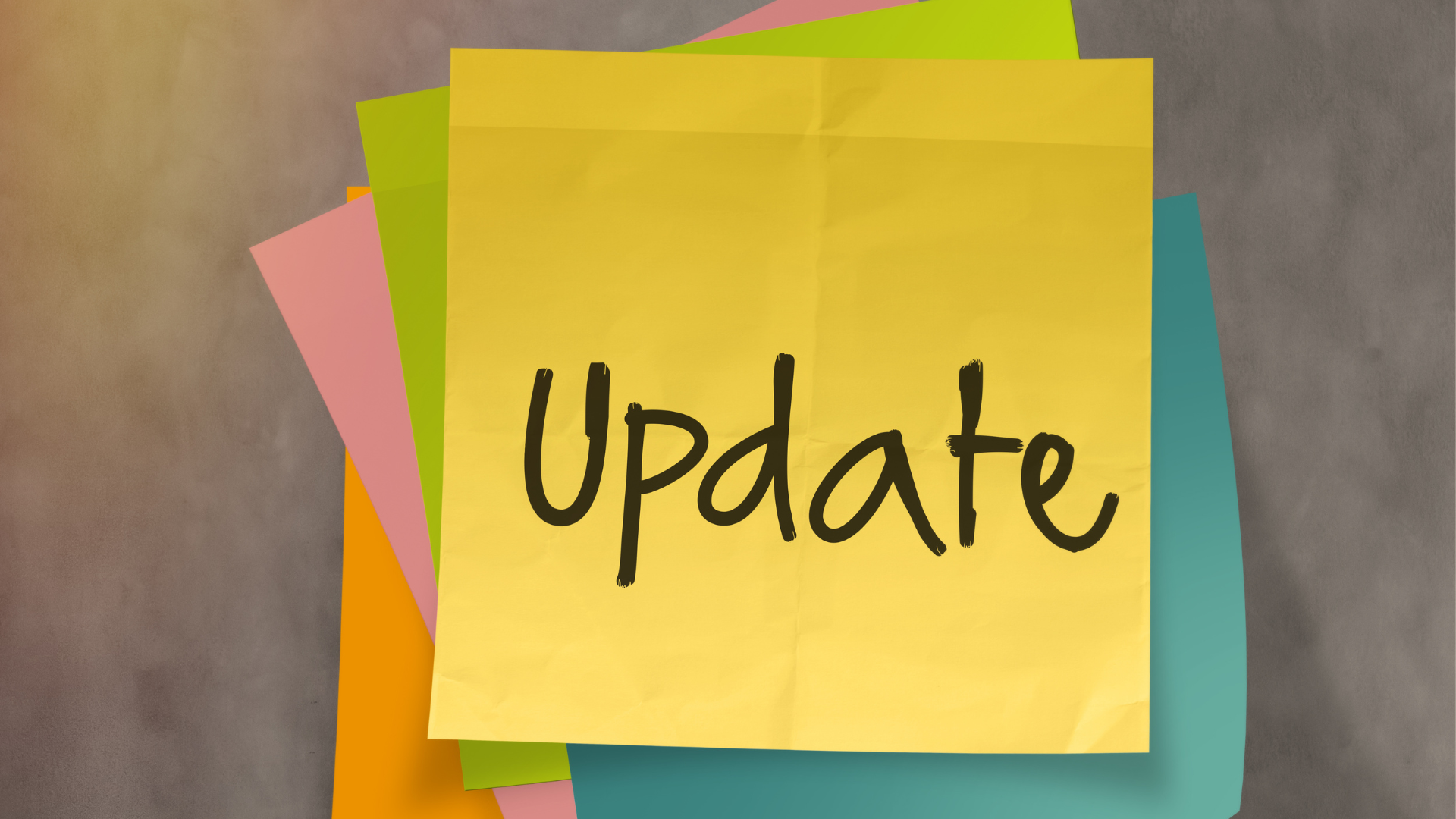 A stack of colorful sticky notes is pinned to a gray surface, with a large yellow note on top displaying the handwritten word "Update" in black ink. 