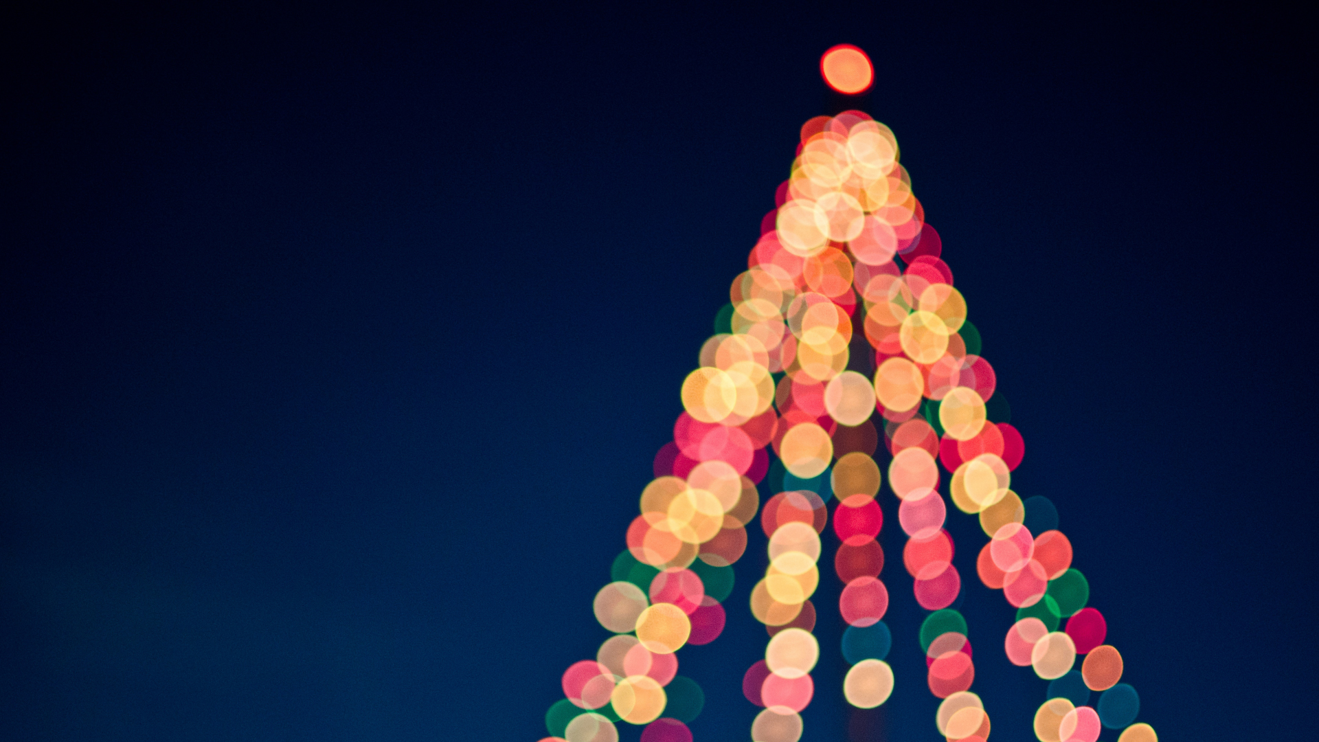 An out-of-focus night time photo of a string light Christmas tree, the red, gold and green lights appearing as bokeh spots forming the pointing tree shape