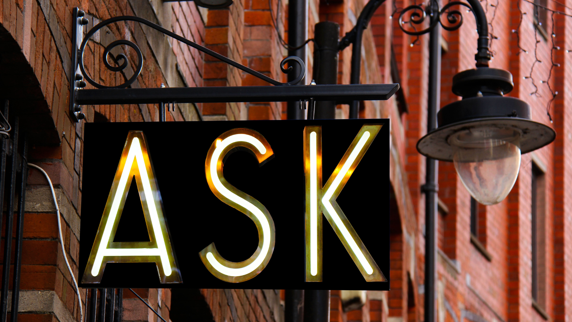 An ornate wrought iron business sign attached to a red brick building with the word "ASK" reflecting in gold
