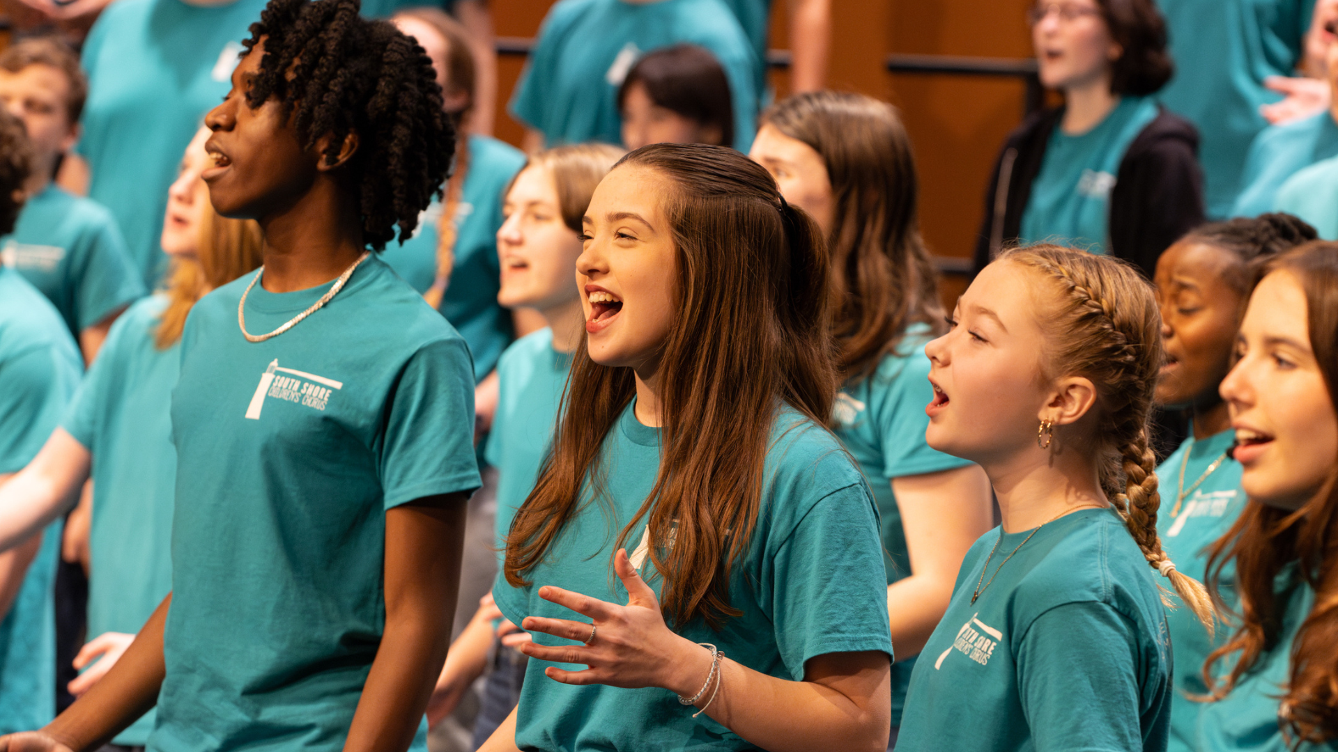 A diverse group of youth smile as they sing in a choir while wearing matching green T-shirts