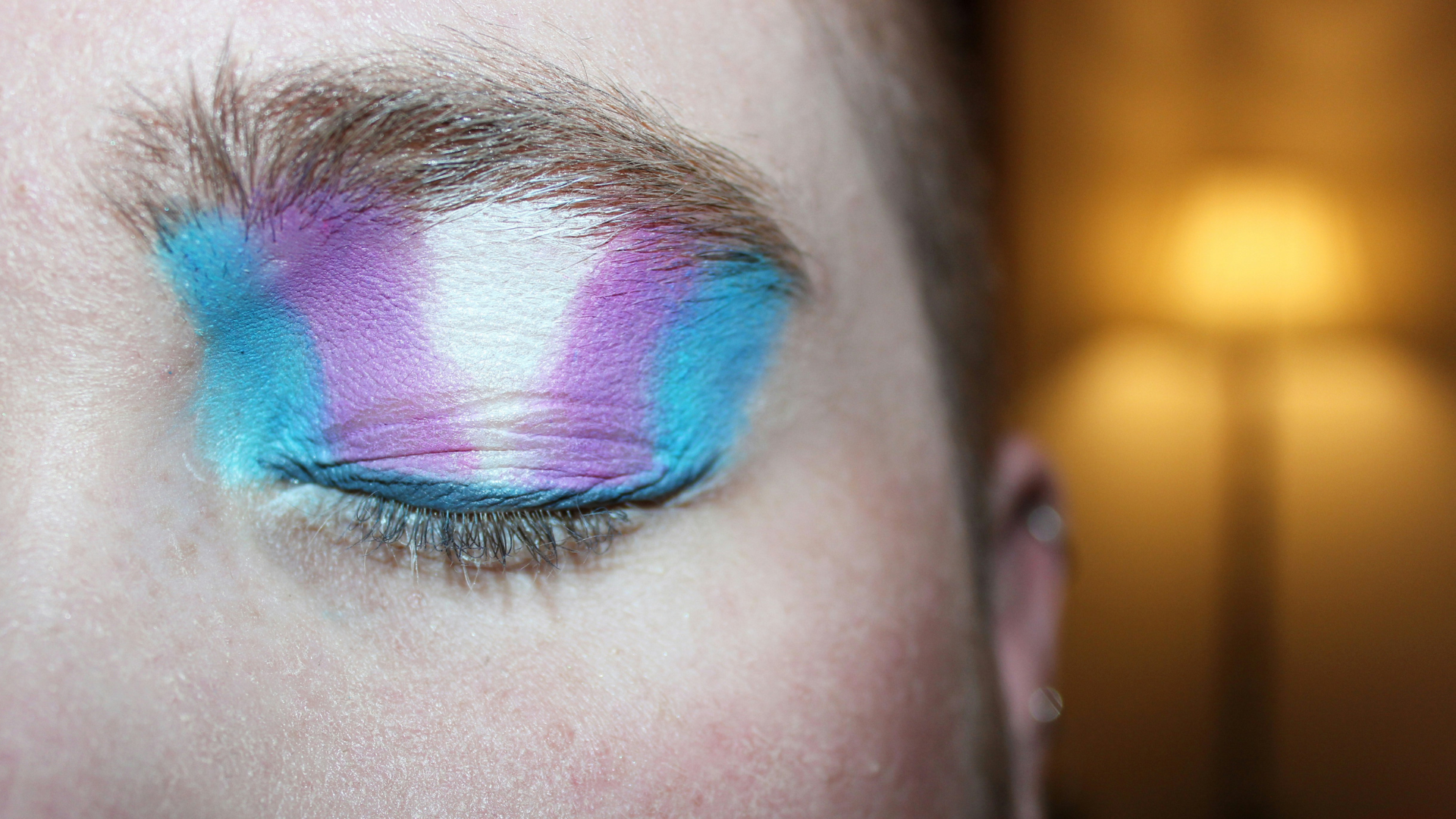 A close up photo of a person's closed eye wearing pink, blue and white eyeshadow with a tall floor lamp glowing in the distance