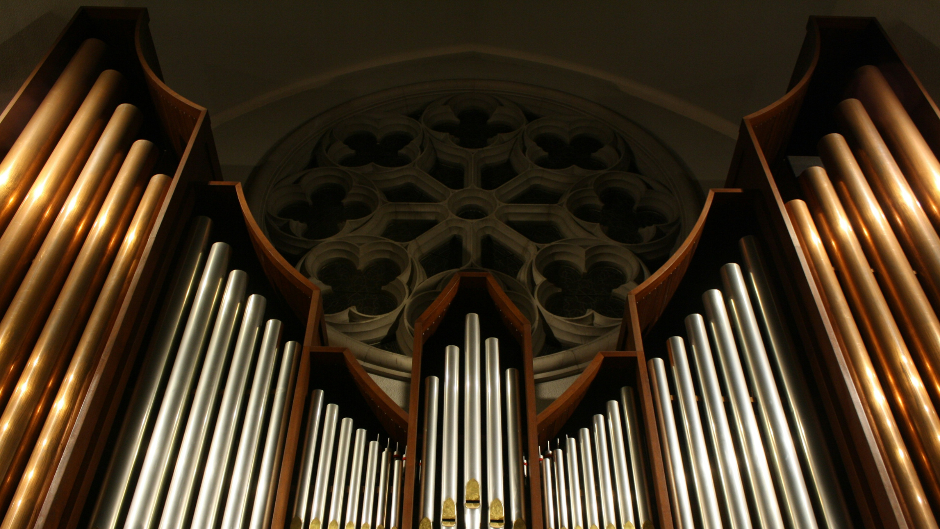 A close view of shiny metal organ pipes, viewed from below, with an intricate carved wood cathedral window in the back