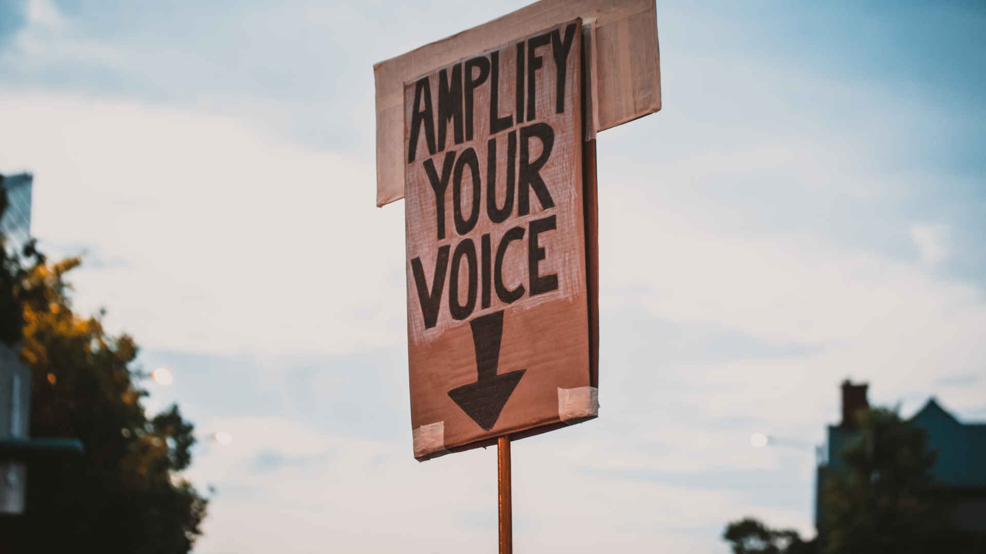A handmade protest sign being held to the sky that reads "Amplify your voice" with an arrow pointing downward