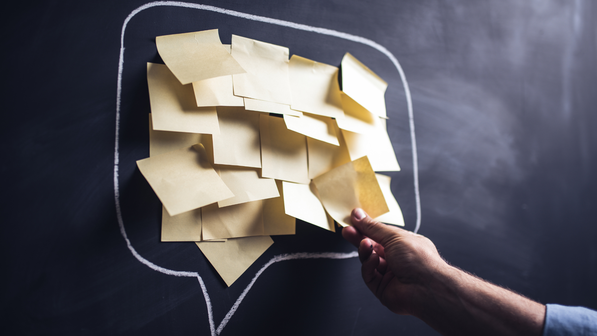 A chalkboard with a speech bubble drawn on it in white chalk; the speech bubble is filled with yellow post-it notes and a hand reaches to add one more to the board