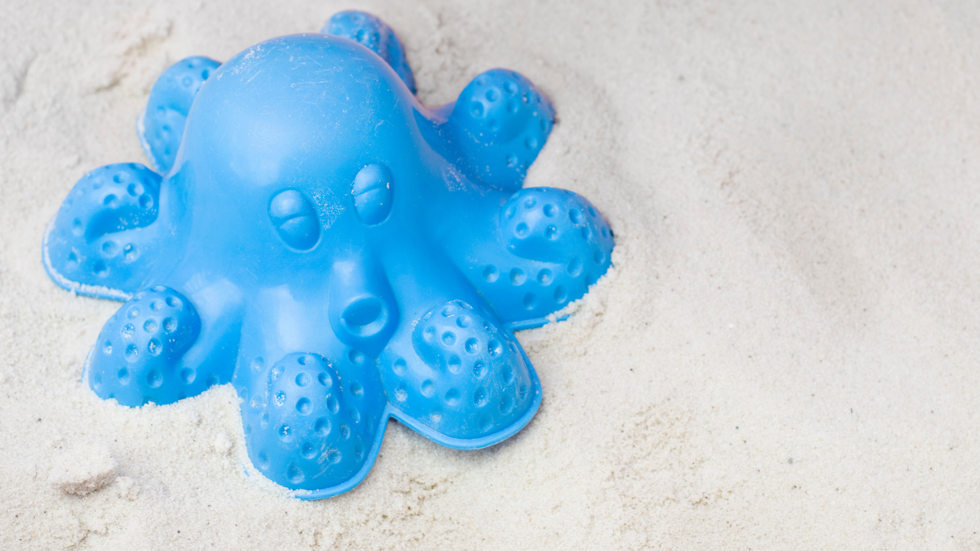 A blue plastic octopus sand castle mold with its mouth in the shape of an "o" sitting on a bed of soft white sand, presumably a beach.
