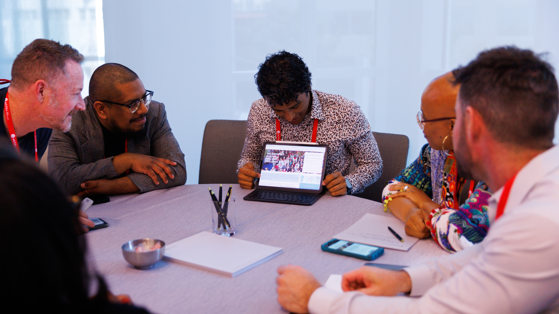 A group of five diverse adults sitting around a table looking toward a man who is holding an iPad with a conference schedule on the screen, facing the camera.