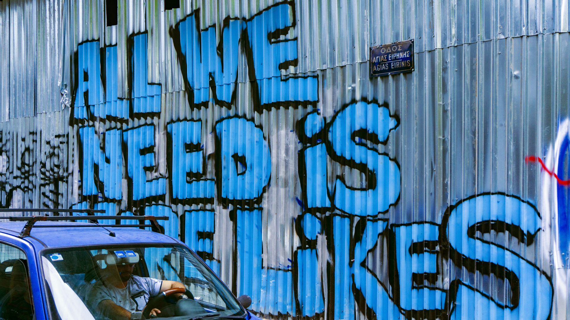 A vehicle parked beside a corrugated metal wall with the words _all we need is more likes_ written on the wall in blue graffiti