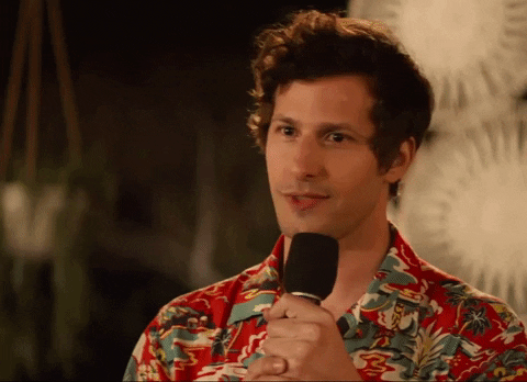Actor Andy Sandberg wears a tropical shirt and holds a microphone to his mouth while mouthing the words "You are not alone."