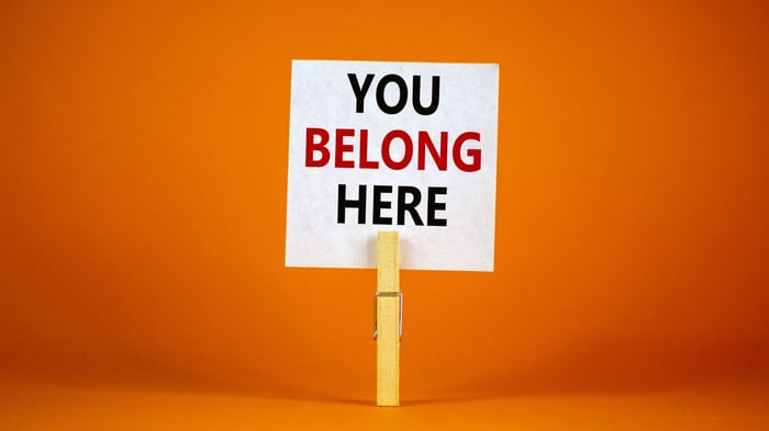 Stock photo with a white square post-it style note with the words "You Belong Here" being held by a wooden clothes peg which is standing upright against an orange backdrop