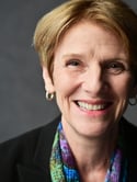 A woman with short blonde hair smiles at the camera while wearing a blue, green and purple scarf and black business blazer - Catherine Dehoney headshot