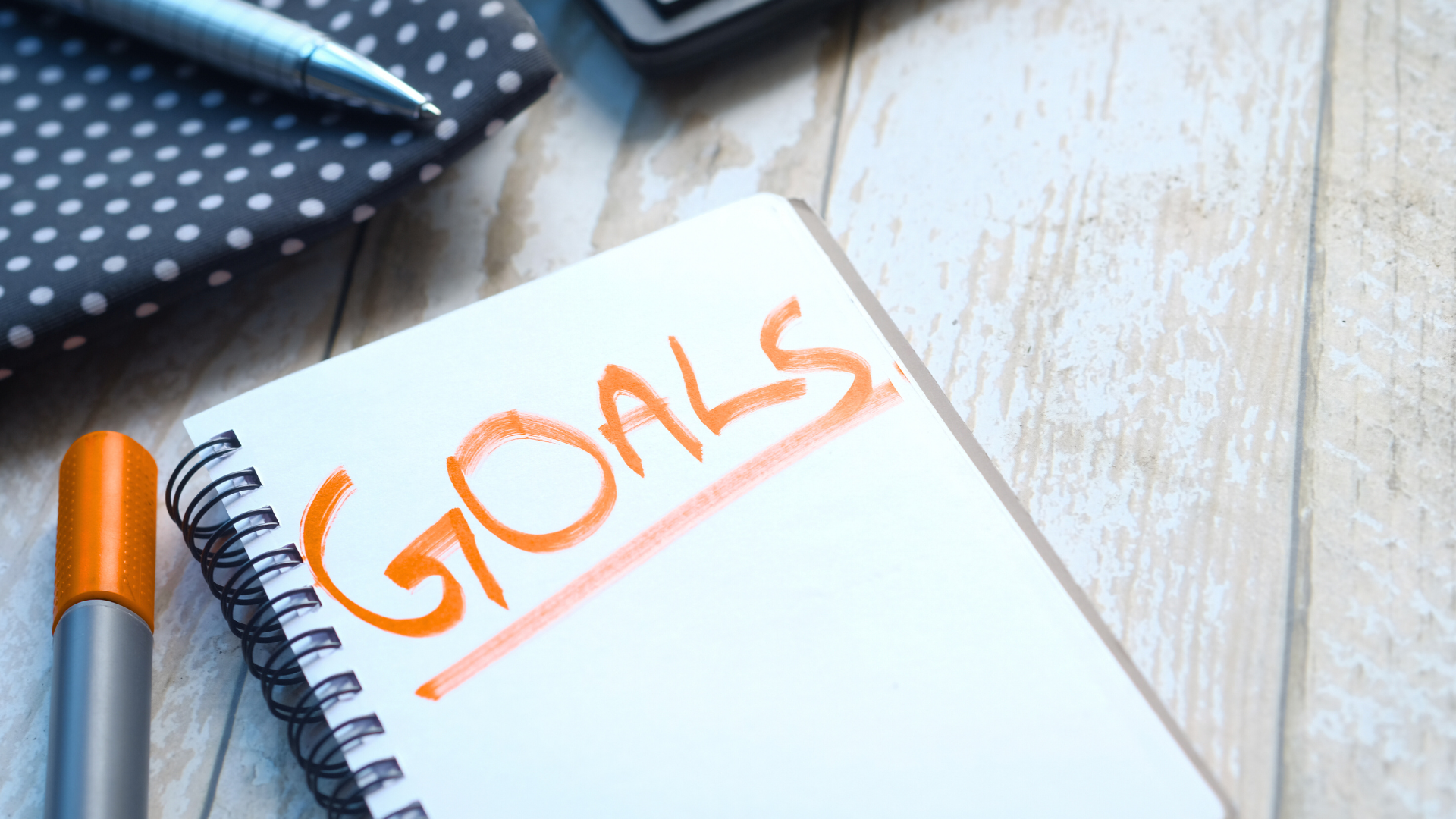 A photo of a spiral-bound notebook open to a page that reads, "GOALS" written with an orange marker which sits to the left slightly out of frame.