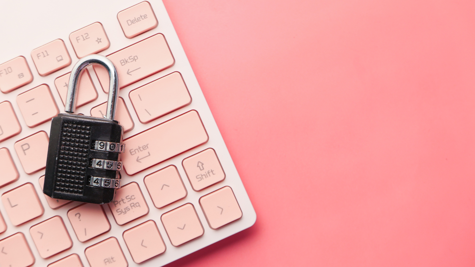 A staged flat lay-style photo of a minimalist-style computer keyboard angled toward the top right corner of image; a black and silver combination lock is positioned on top of the keys alluding to the concept of digital security