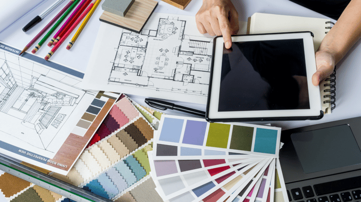 Top down view of an interior designer's desk which is covered with floor plans, color palettes and hands holding a tablet