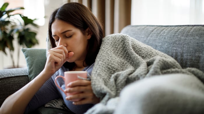 Photo of a woman curled up on a sofa covered in a cozy blanket, holding a mug while coughing into her other hand