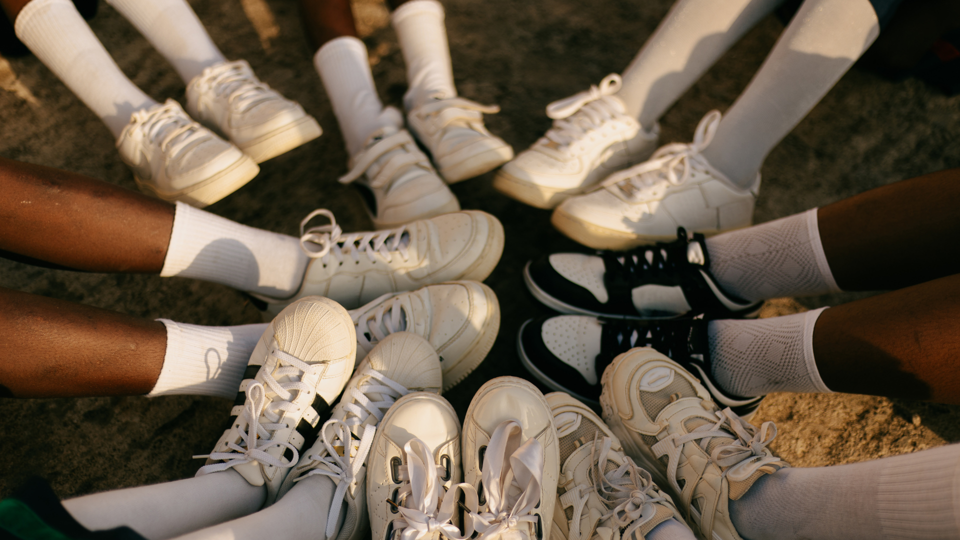 A cropped photograph of youth sitting on the ground in a circle, all wearing sneakers and white socks, with their shoes touching in the center of the circle