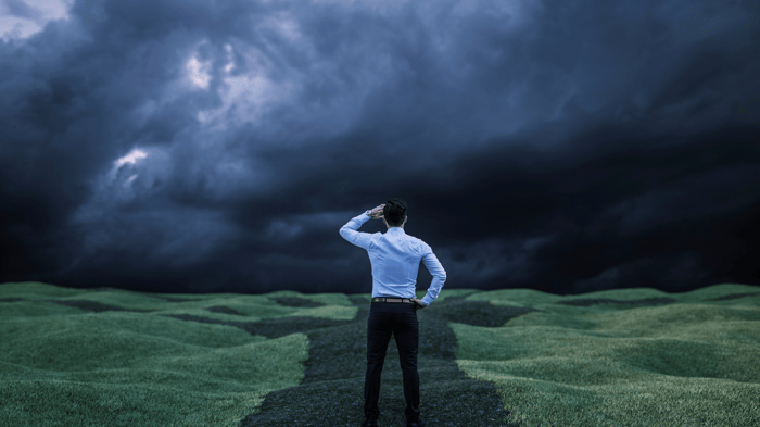 The image features a person standing in a vast, grassy landscape with a stormy sky above. The individual is wearing a light blue shirt and dark pants and is facing the ominous clouds. They are saluting or shielding their eyes with one hand, while the other hand rests on their hip. The scene conveys a sense of contemplation or facing challenges. The atmosphere is dramatic with the dark clouds indicating an impending storm.