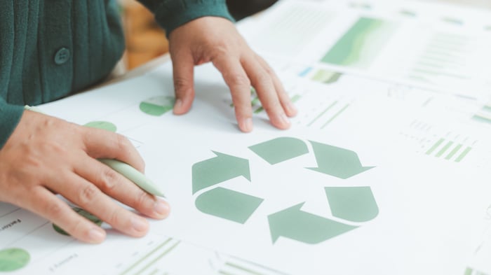 Photo of a person's hands holding a pen as their hands touch a piece of paper with a green recycle symbol printed on it