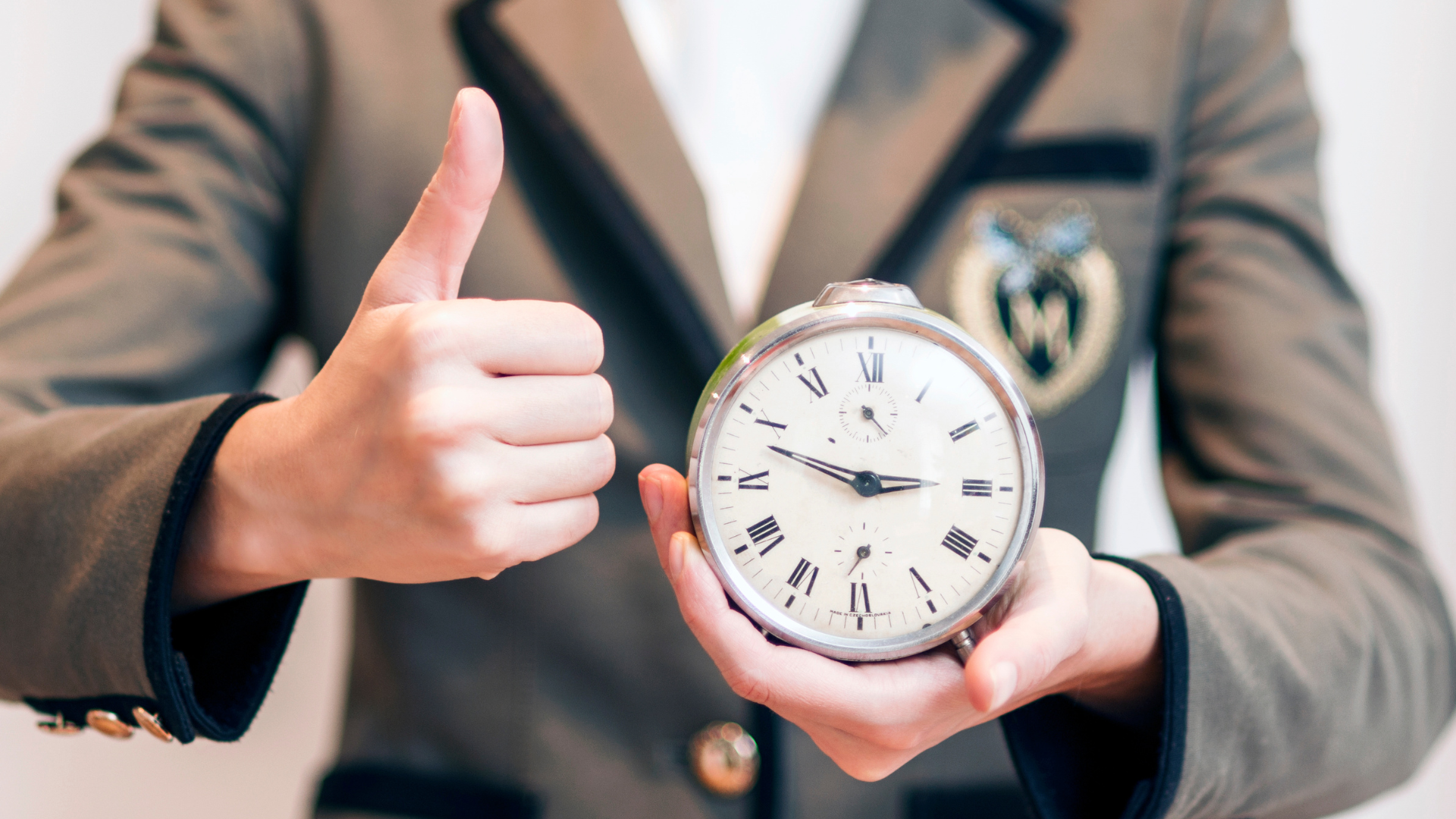 A photo of a person in a blazer holding clock in one hand and giving a thumbs up with the other