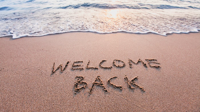 An ocean wave washes up on a shore at sunset with the words "Welcome Back" inscribed in the sand