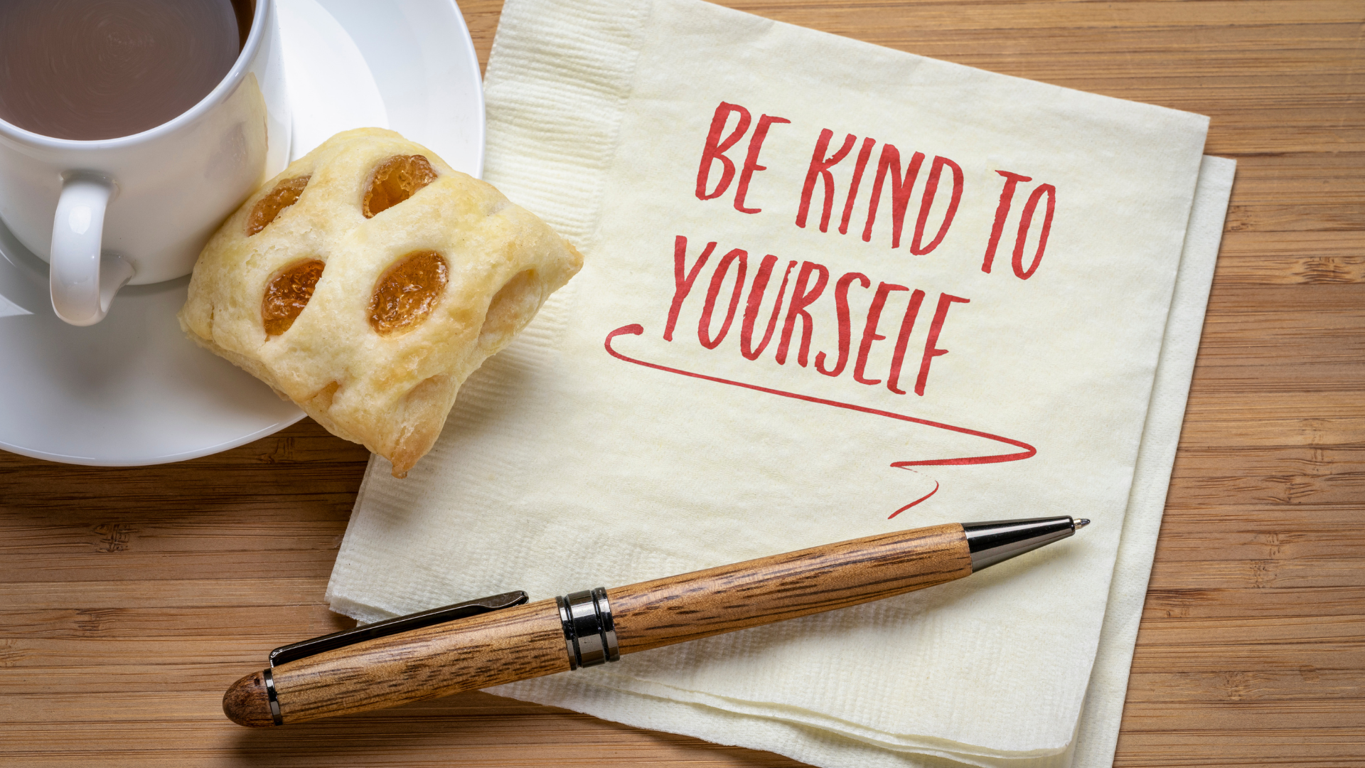 Stock photo of a napkin next to a coffee cup and danish, a pen lays across the napkin and handwritten message "Be Kind to Yourself" is written on the napkin.