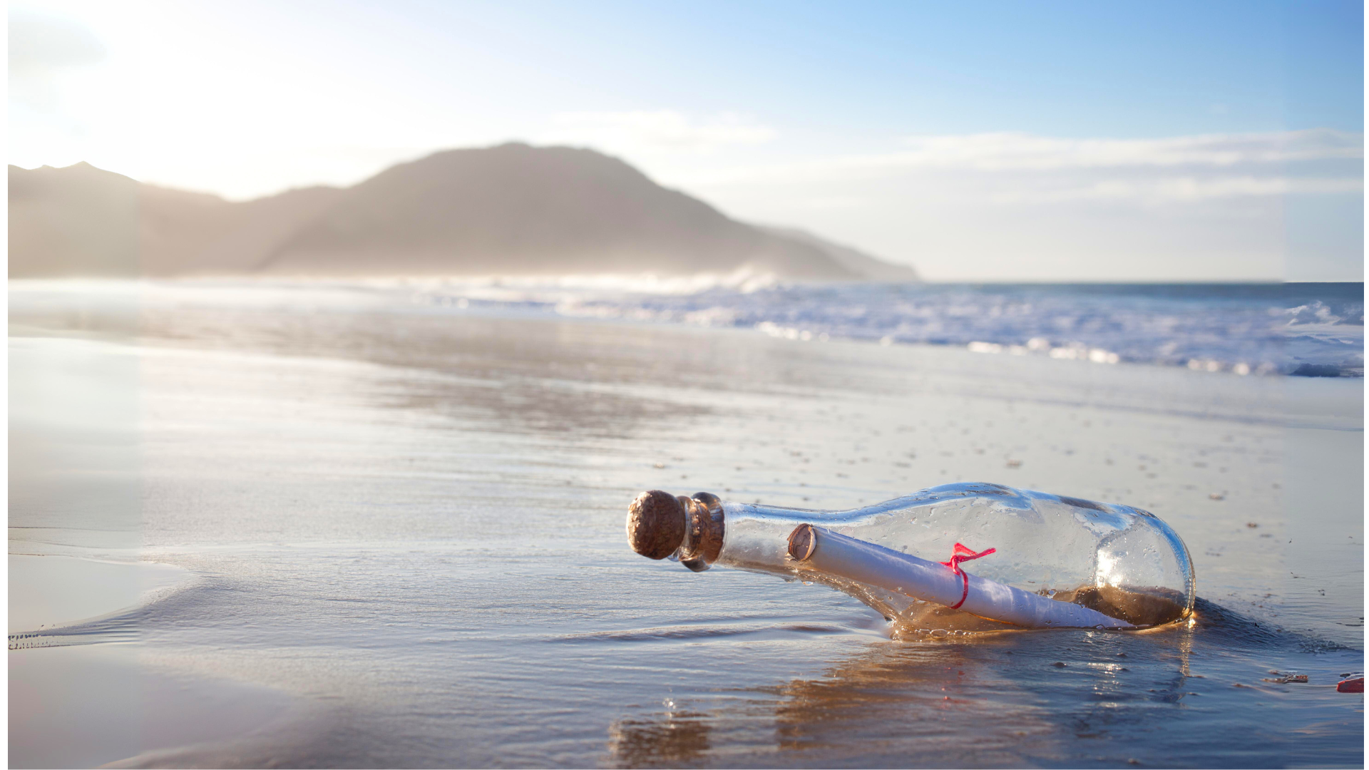 A clear glass bottle washed up on a sandy beach with a white paper note rolled up inside and tied with a red string. In the distance waves wash ashore and a radiant, cool blue sky is partly hidden behind a mountain that appears on the horizon.
