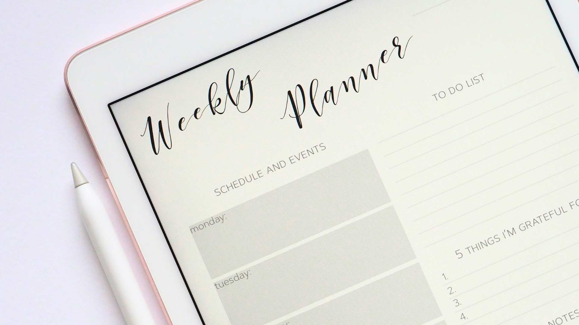 Flat lay photograph of a turned-on silver iPad beside displaying a weekly planner with an Apple Pencil sitting to the left