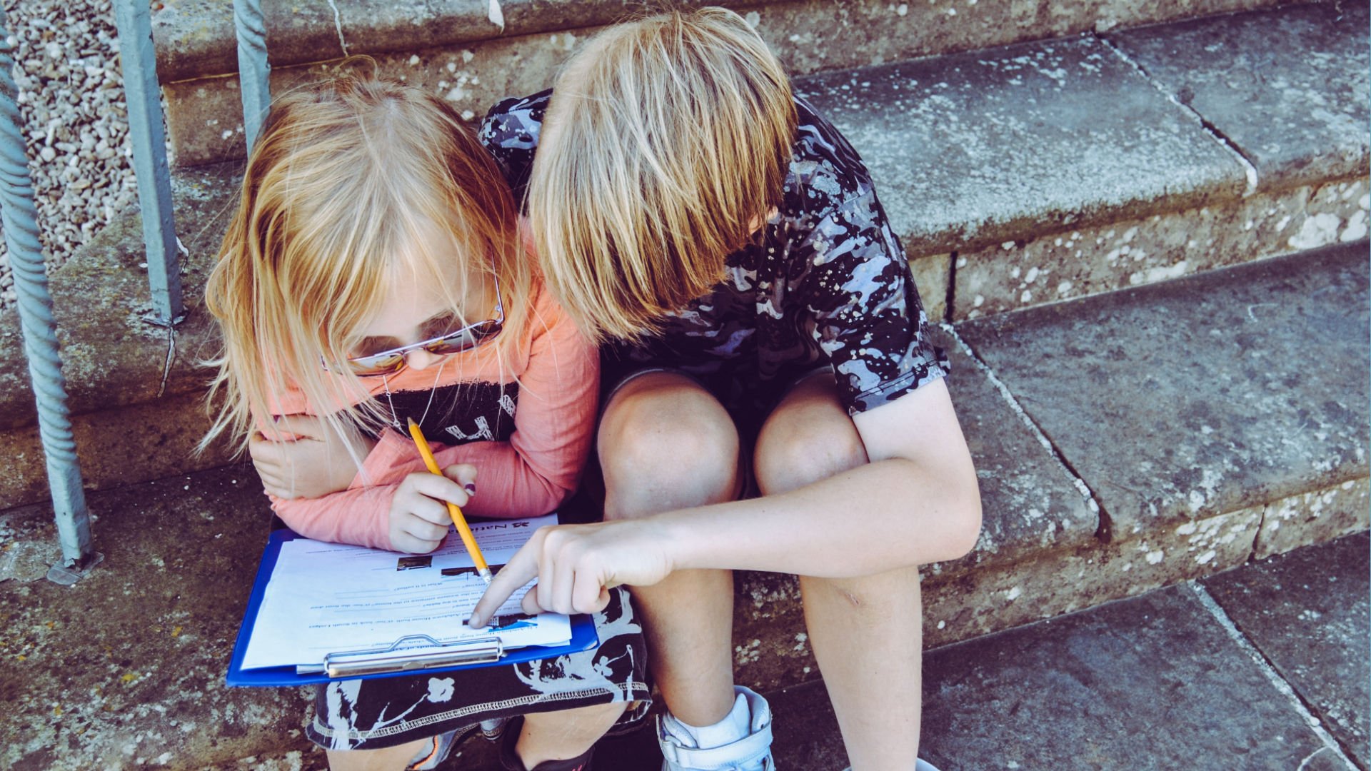 Two children sitting outside on stairs, working together on a document.