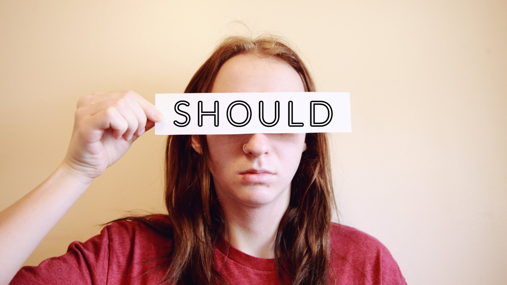 A person holding a narrow white piece of paper that covers only their eyes and the paper says "SHOULD" on it.