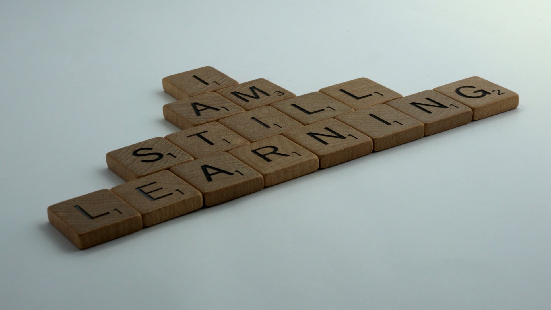 Scrabble tiles lying flat on a white background that spell out "I Am Still Learning"