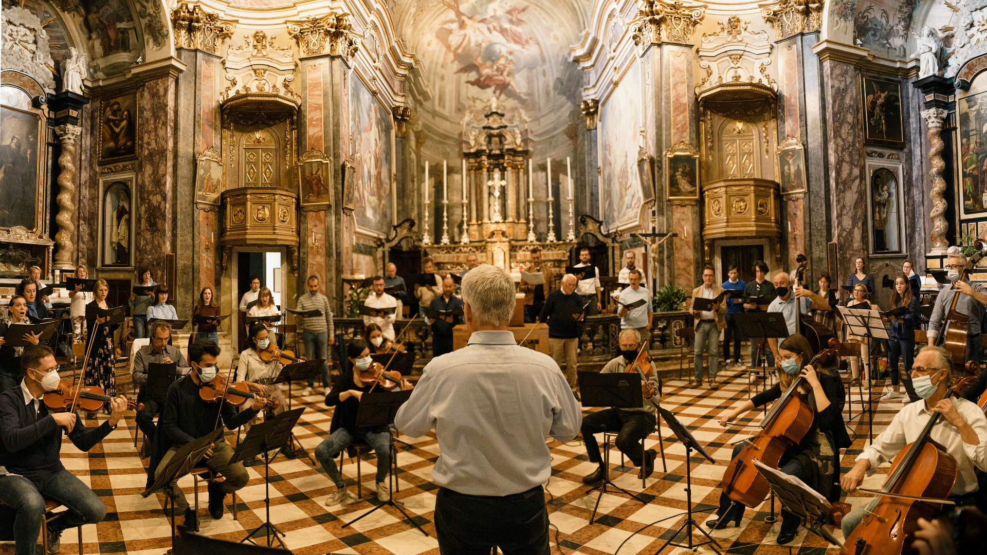 Members of an orchestra sitting on chairs in front of a chamber choir inside an ornate cathedral