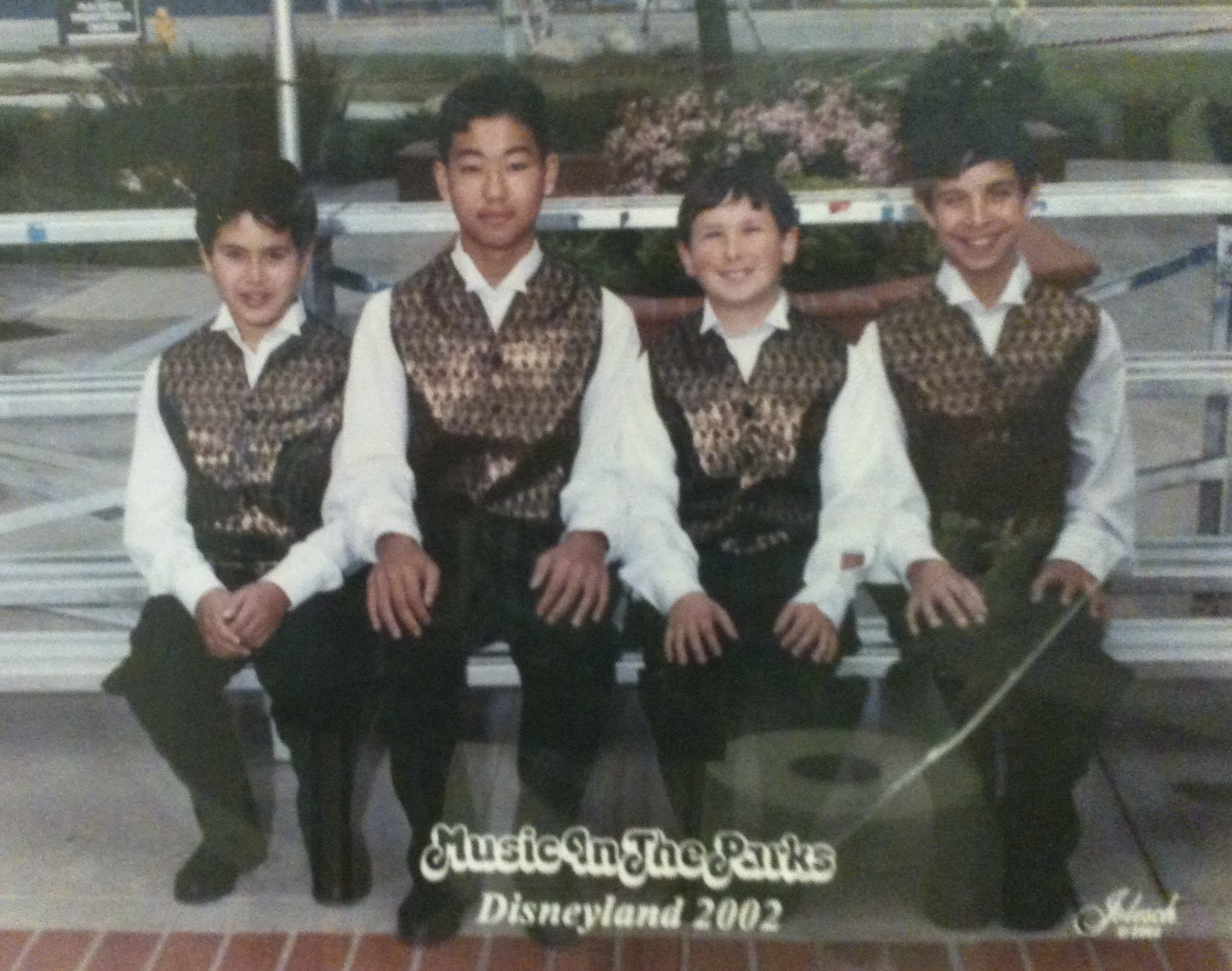 A grainy photo of four clean-cut boys sitting side by side on a bench, facing the camera, wearing white button collard shirts, black pants and fancy vests. The boy on the far left is Chorus Connection founder Jacob Levine when he was in 7th grade. The photo bears text that reads "Music In The Parks - Disneyland 2002"