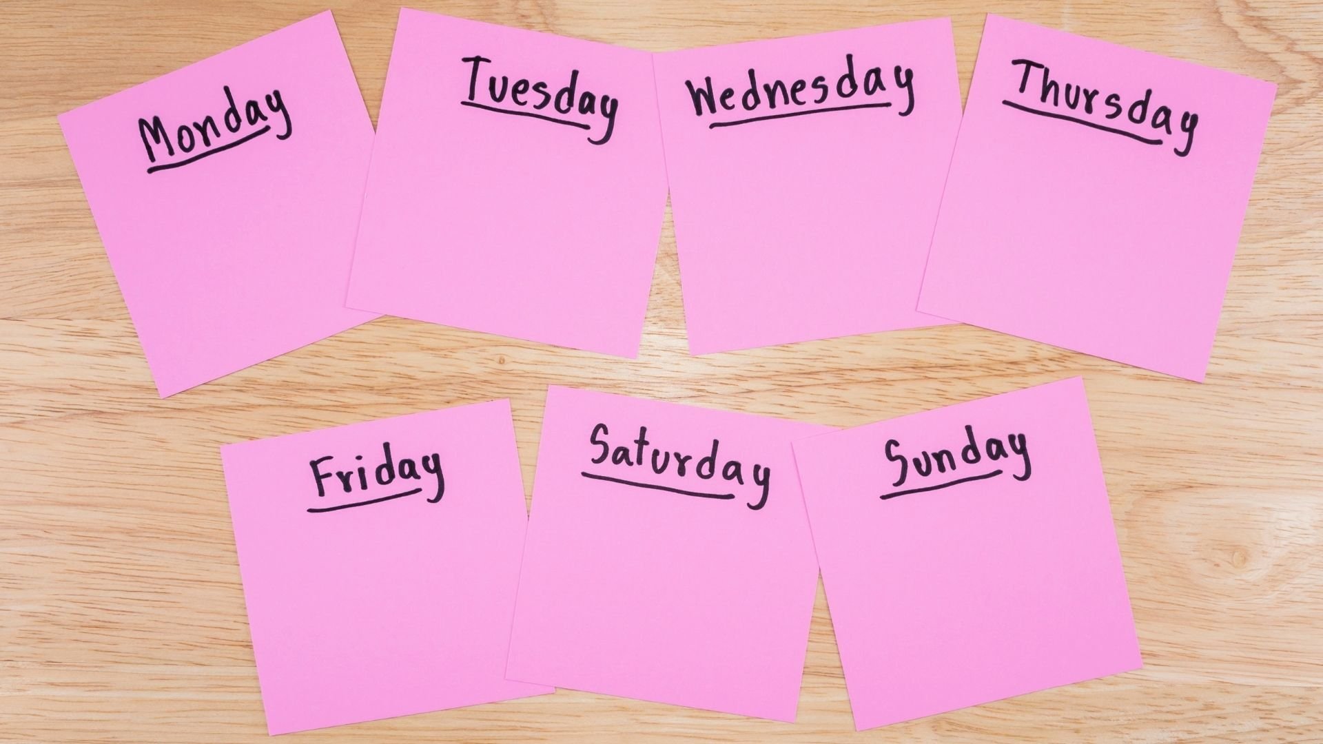 seven pink sticky notes with a day of the week written and underlined in bold black handwriting stuck to a wood grain surface