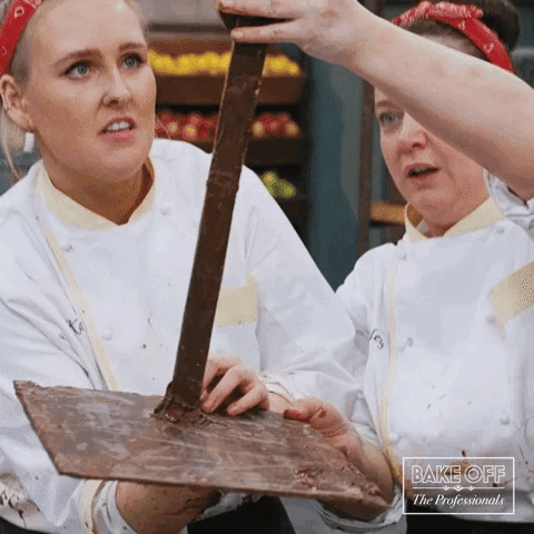 Two women in a kitchen trying to stand a chocolate tower up straight, but it tips and they both fumble as they try to catch it