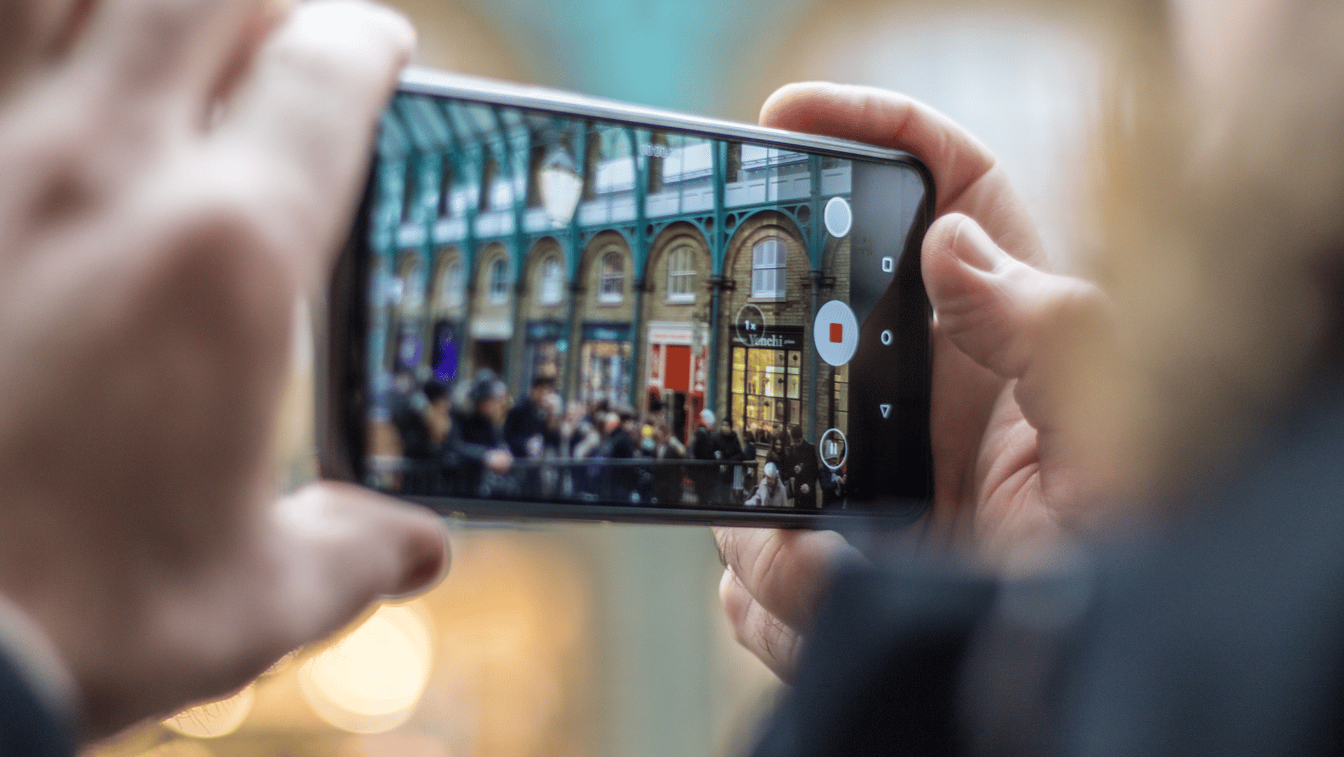 Close-up of a person holding a smartphone horizontally while recording a video of a crowded urban street scene. The phone screen shows colorful storefronts, arched windows, and a group of people in the frame.