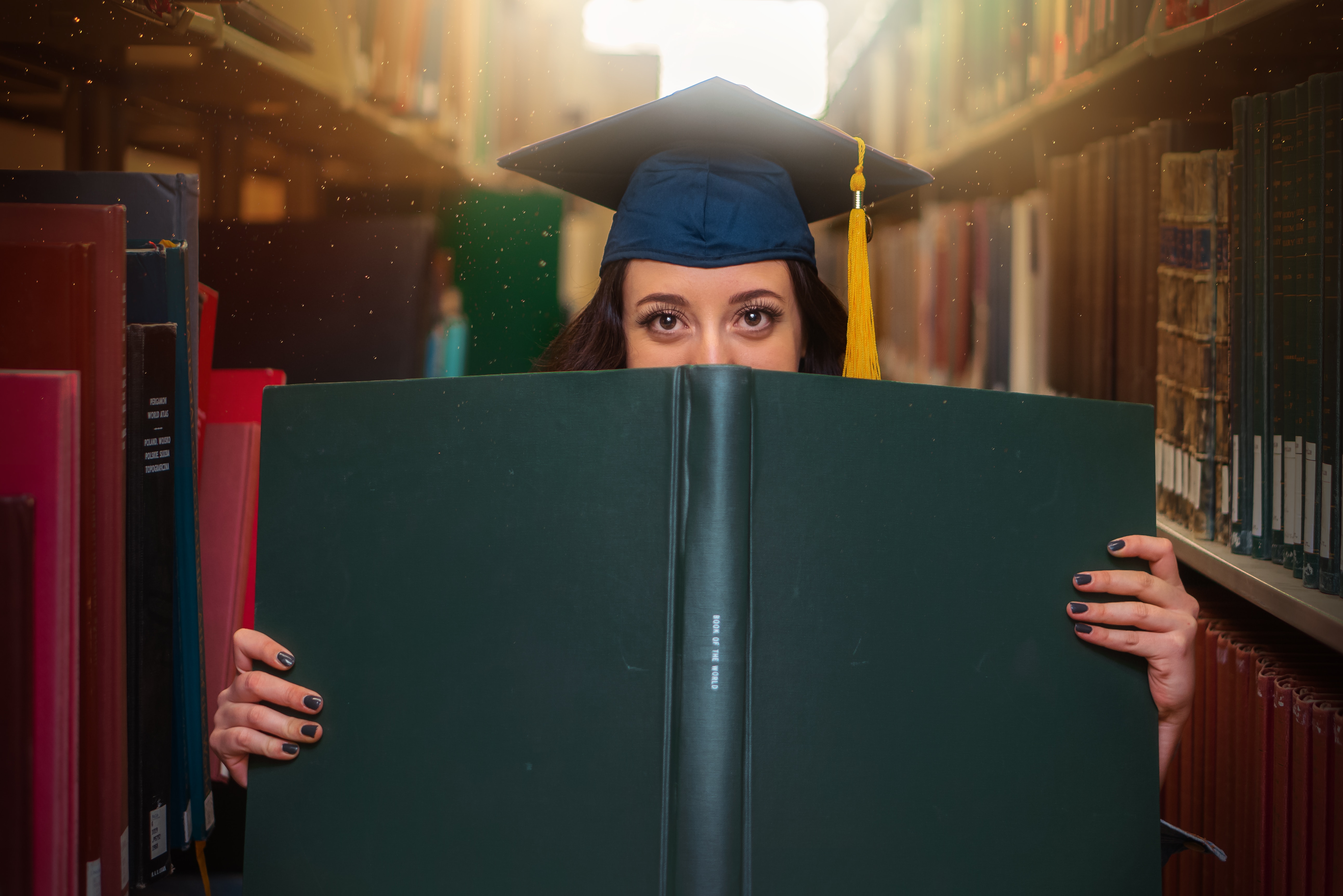 A young woman with brown hair, brown eyes and long eye lashes holds open a large book in front of her face, covering her nose, mouth and chin. She's standing in 'the stacks' of a library, surrounded by rows of books and she is wearing a graduation cap. The sun glows in through the window behind her.